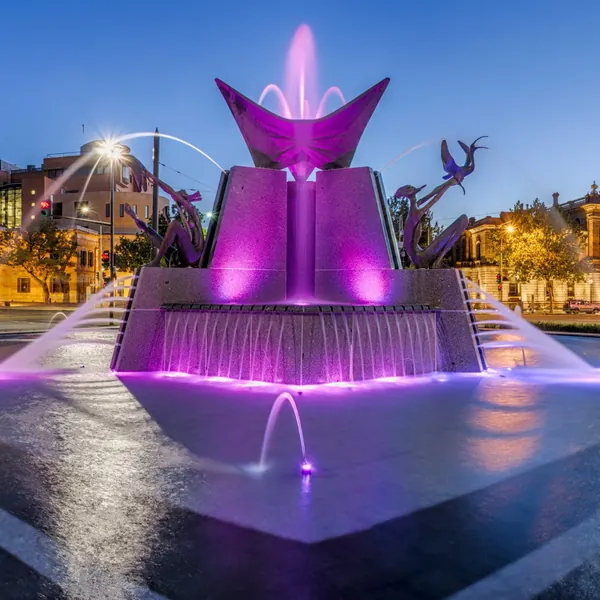 The Victoria Square fountain at dusk, lit violet, water falling from its tiers onto wet paving